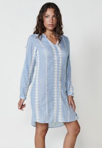 Helles blaues Kleid mit langen Ärmeln und V-Ausschnitt. Verfügt über vertikale weiße Stickmuster und einen leicht abgerundeten Saum. Hergestellt aus leichtem Stoff.