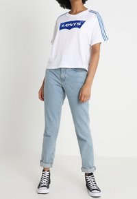 Camiseta blanca corta con logo azul de Levi's, con franjas azules en los hombros; combinada con vaqueros de denim de color azul claro y zapatillas altas negras.