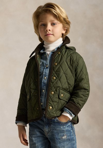 Joven niño con chaqueta acolchada verde oliva, camisa vaquera, jersey de cuello alto blanco y vaqueros azules, de pie con una mano en el bolsillo.
