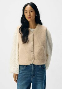 Fleece vest in lichtbeige met een zachte textuur, ronde halslijn en drukknoopsluiting, gedragen over een witte, met patroon bedrukte lange mouw top.