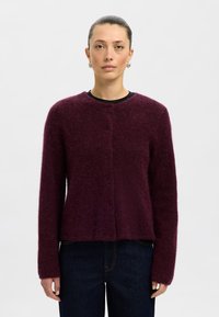 Vinröd kofta tillverkad av mjukt, luddigt material. Har en rund halsringning, långa ärmar och knappstängning fram.