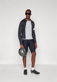Fahrradausrüstung bestehend aus einer schwarzen Langarmjacke, einem grauen Basisschichtoberteil, schwarzen Shorts, schwarzen Schuhen und einem grauen Helm, mit Sonnenbrille.