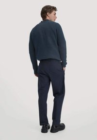 Marineblauwe geribbelde gebreide trui met een ontspannen pasvorm, gecombineerd met donkere broeken en zwarte schoenen, gezien van achteren.