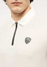 Λευκό πόλο πουκάμισο με φερμουάρ στον λαιμό, με μαύρο λογότυπο EA7 στη θώρακα. Ομαλή υφή, μοντέρνο σχέδιο και λεπτομέρειες σε αντίθεση.