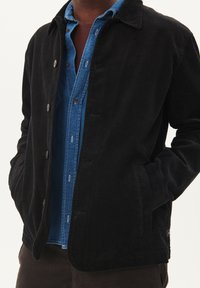 Black corduroy jacket with a collared neckline, front button closure, and two side pockets, worn over a blue denim shirt.