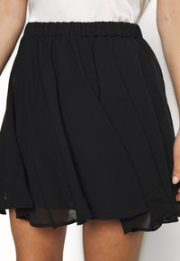 Musta hame, jossa on kerätty joustava vyötärö, valuva kangas ja kerroksellinen muotoilu. Siinä on lyhyt, levenevä malli ja läpikuultava helma.