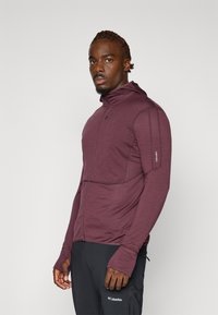 Bordeaux-farvet zip-up jakke med høj krave, syet detaljer og tommelfingerhuller ved ærmerne, parret med sorte bukser med et logo på lommen.