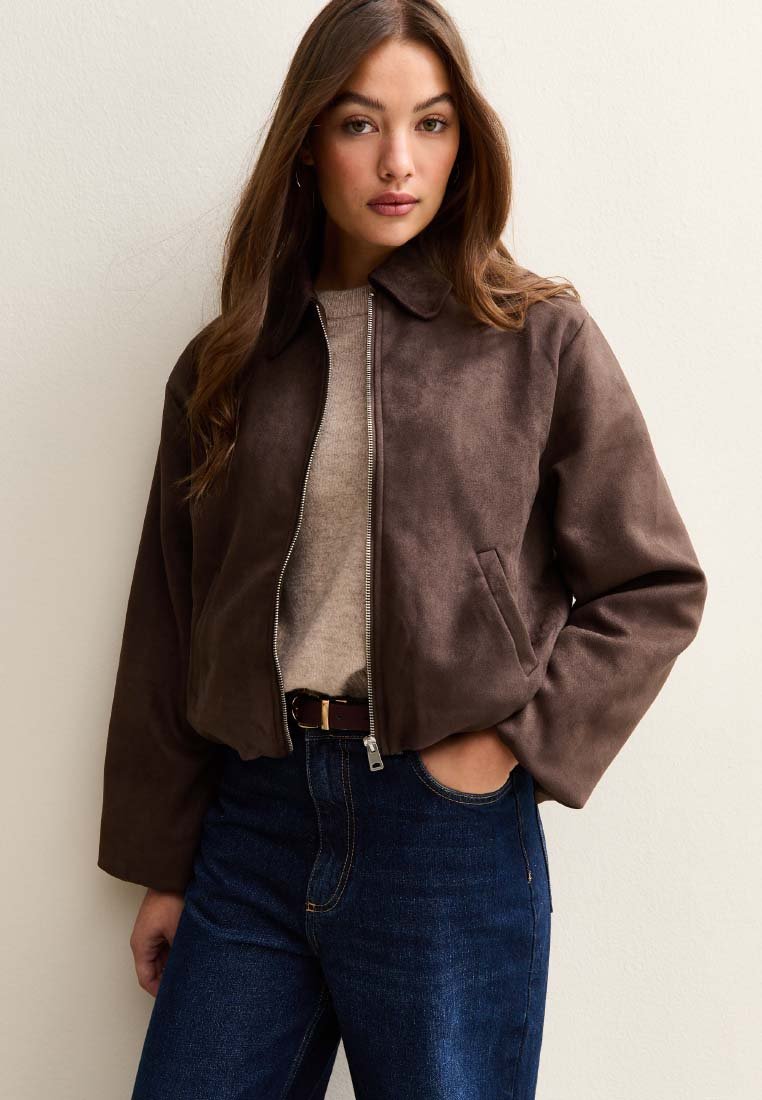 Veste courte en daim marron avec une fermeture éclair, un col basique et des poches latérales, portée sur un pull beige et associée à un jean en denim foncé.