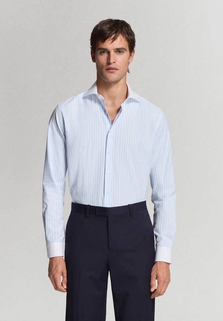 Jeune homme portant une chemise à rayures bleu clair et un pantalon bleu marine foncé, debout devant un fond gris uni.