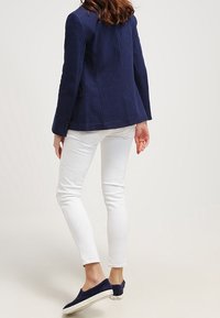 Blazer ajusté bleu marine au design structuré, associé à un jean blanc ajusté. Les chaussures sont des mocassins bleu foncé avec une semelle blanche.