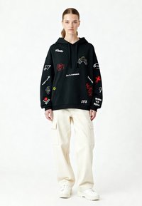 Felpa nera con diverse grafiche ricamate colorate, tra cui testo e simboli. Abbinata a pantaloni cargo color crema e sneakers bianche.