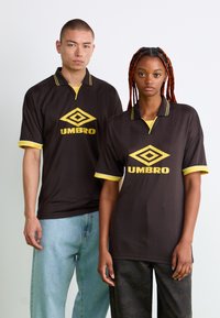 Hnedé tričká s krátkymi rukávmi so žltými akcentmi a geometrickým vzorom. Obsahujú logo Umbro a dizajn polo goliera.