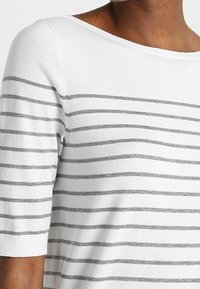 Bílý top se šedými vodorovnými pruhy, s širokým výstřihem a krátkými rukávy. Materiál působí měkce a má hladkou texturu.