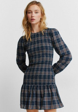 Vaaleahiuksinen nainen, jolla on irtonaiset hiukset ja päällään istuva tummansininen ruudullinen mekko, jossa on pitkät pöyhistetyt hihat ja röyhelöinen helma, seisoo käsivarret selän takana.