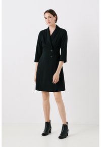Robe noire à la coupe ajustée, boutons double boutonnage, col châle et manches trois-quarts, associée à des bottines à la cheville.