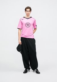 Ροζ αθλητική φανέλα με μαύρες λεπτομέρειες, που διαθέτει στρογγυλό λογότυπο με το "90". Χαλαρές μαύρες παντελόνες, μαύρο καπέλο στο χέρι και μαύρα αθλητικά παπούτσια.