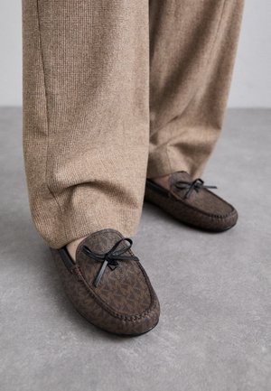 Person iført brune teksturerede loafers med sorte sløjfedetaljer kombineret med løse beige ribstrikkede bukser på gråt gulv.