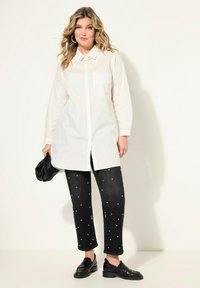 Hvid langærmet skjorte med knapper og krave samt brystlomme, parret med sorte jeans prydet med perle-lignende accenter og sorte loafers.