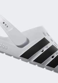 Biele sklzovací sandál vyrobený z gumy s troma čiernymi pruhmi, nastaviteľným popruhom a ventilačnými otvormi pre priedušnosť, so značkou Adidas.