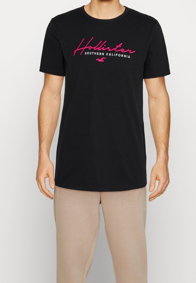 Svart bomulls t-shirt med korta ärmar, med rosa broderad text "Hollister" och "Southern California" centrerad på bröstet.