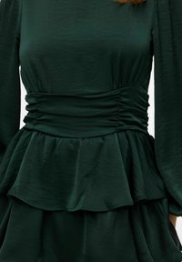 Tumši zaļa kleita ar savāktu korseti, pepluma jostasvietu un plūstošām kārtām. Audumam ir gluda tekstūra ar smalku spīdumu.