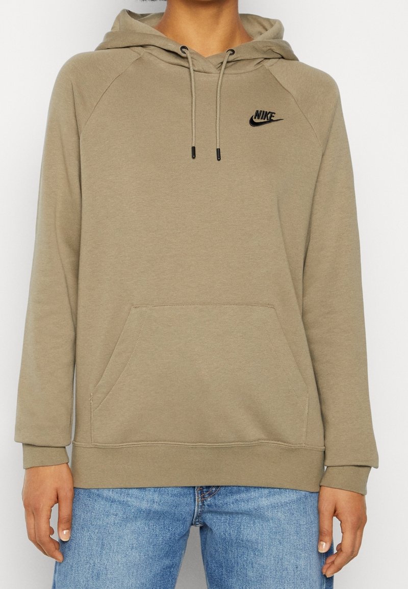 Personne portant un sweat à capuche beige Nike avec poche avant et jean bleu, cadré au niveau du cou et du milieu de la cuisse.