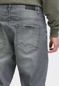 Pelēkas džinsa bikses ar šauru piegriezumu un aizmugures kabatām, uz kurām redzams zigzaga dūriena raksts. Vidukļa daļā ir melnas etiķetes detaļa.