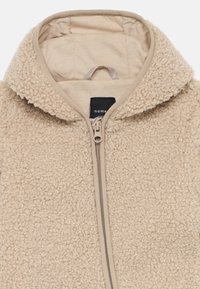 Beige Fleecejacke mit einer strukturierten, flauschigen Außenseite, einem Reißverschluss vorne und einer gemütlichen Kapuze. Das Etikett ist schwarz mit weißem Text.
