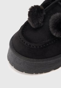 Chausson en suède noir avec deux pompons moelleux sur le dessus. Semelle en caoutchouc texturé épais avec un design superposé autour du bord.
