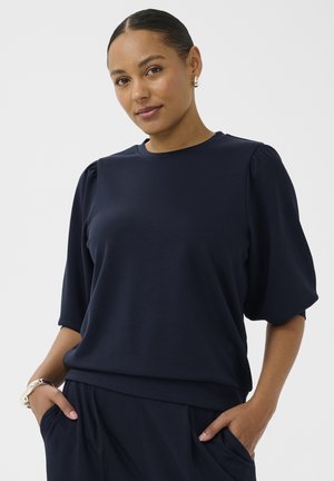 Kobieta z ciemnymi włosami zaczesanymi do tyłu, ubrana w granatową bluzkę z bufkami i dopasowane spodnie, ręce w kieszeniach, na jednolitym jasnym tle.