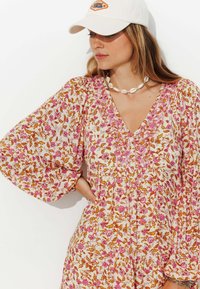 Robe florale avec une base blanche et des motifs roses et oranges, un col en V, des manches bouffantes et des détails élaborés. Associée à une casquette blanche.