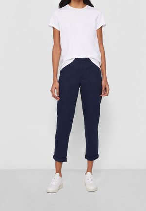 Femme portant un t-shirt blanc à manches courtes, un pantalon bleu marine retroussé et des baskets blanches, debout devant un fond uni gris clair.