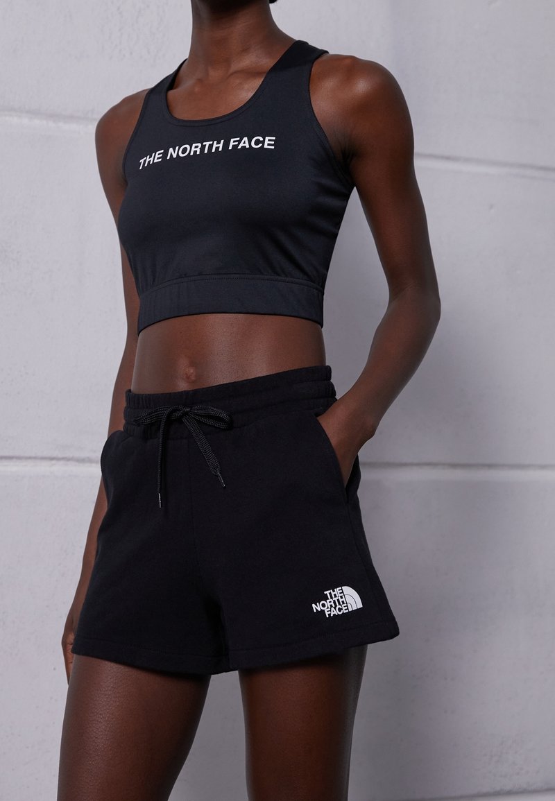 Femme portant un crop top noir sans manches et un short noir, tous deux avec le logo "The North Face", debout contre un mur gris clair.