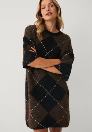 Kvinde med lyst hår iført en brun og sort argyle-mønstret sweaterkjole med armene krydset mod en ensfarvet baggrund.