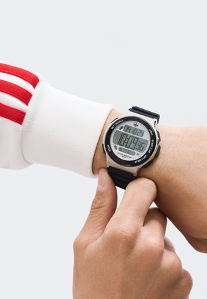 Digitaalne kell musta kummirihmaga ja hõbedase korpusega. Ekraan näitab aega, kuupäeva ning sellel on nupud režiimi, valguse ja meenutamise jaoks.