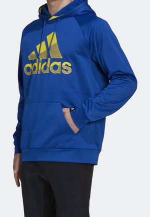 Homme portant un sweat à capuche Adidas bleu avec logo jaune, main dans la poche avant, et pantalon noir sur fond uni.