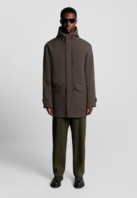 Bruine waterdichte jas met een hoge kraag, twee voorzakken en een ritsclosure. Gecombineerd met olijfgroene broek en zwarte schoenen.