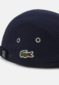Căciulă marină din material texturat, cu ochiuri din metal argintiu și un logo brodat de crocodil verde și roșu în față. Curea ajustabilă la spate.
