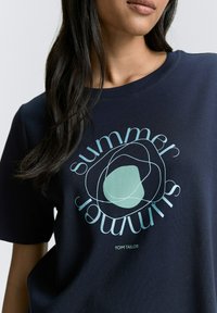 Tamsiai mėlynas medvilninis marškinėliai su apvaliu kaklu, išsiskiriantys šviesiai mėlynu abstrakčiu dizainu ir žodžiu "vasara" meniniu šriftu. Prekės ženklas: Tom Tailor.