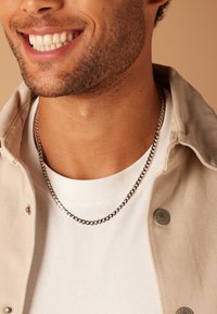 Zilveren ketting gedragen over een witte shirt, met een beige jas. De ketting heeft in elkaar grijpende schakels en heeft een gladde, glanzende afwerking.