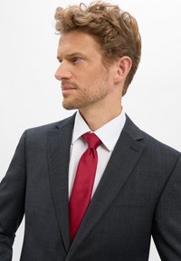 Granatowa teksturowana marynarka z pojedynczą kieszenią, biała koszula formalna i gładki czerwony jedwabny krawat. Widok z profilu, krótkie falujące włosy.