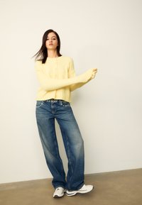 Cardigan en maille jaune clair avec boutons sur le devant et manches longues, associé à un jean bleu ample et des baskets blanches. Texture lisse et coupe décontractée.