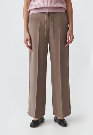 Bequeme Hose im taupefarbenen Stoff mit einem Kordelzugbund und klaren Falten. Kombiniert mit einem hellrosa gerippten Strickoberteil und schwarzen Ballerinas.