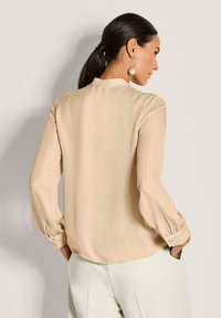 Beige Bluse mit hohem Kragen und langen, puffigen Ärmeln. Glatter Stoff mit lockerer Passform, ausgestattet mit Knopfmanschetten und einem dezenten Glanz.
