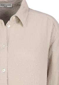 Hellbeiges Hemd zum Zuknöpfen aus strukturiertem Stoff mit kleinen, erhabenen Punkten, ausgestattet mit spitzem Kragen und weißen Knöpfen.