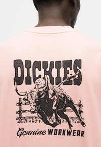 Άτομο που φοράει ανοιχτό ροζ πουκάμισο με μαύρο γραφικό ενός ταύρου που τρέχει μπροστά από ξύλινο φράχτη και κείμενο που γράφει "DICKIES Genuine WORKWEAR."