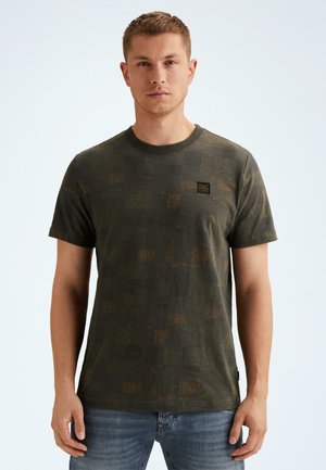 Man draagt een donkergroene t-shirt met korte mouwen en patroon, en een blauwe spijkerbroek, staand tegen een effen lichte achtergrond.