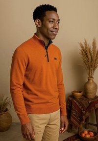 Oranje sweater met rits en structuur breiwerk, gecombineerd met beige broek. Op de achtergrond zijn decoratieve aardewerkstukken en planten te zien, met een patroon van stof.