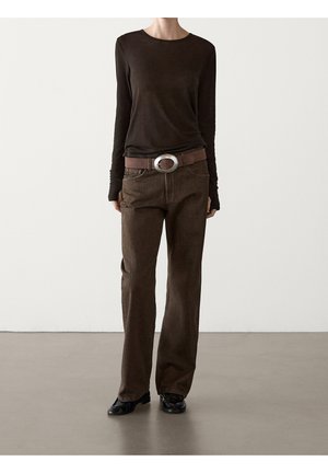 Persona che indossa una maglia a maniche lunghe marrone scuro, pantaloni marroni a gamba dritta, cintura marrone con grande fibbia rotonda e scarpe nere, in piedi su un pavimento grigio.