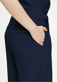 Námořnické modré kalhoty s hladkým povrchem, s boční kapsou a malou značkovou cedulkou. Tkanina vypadá lehce a dobře padnoucí.